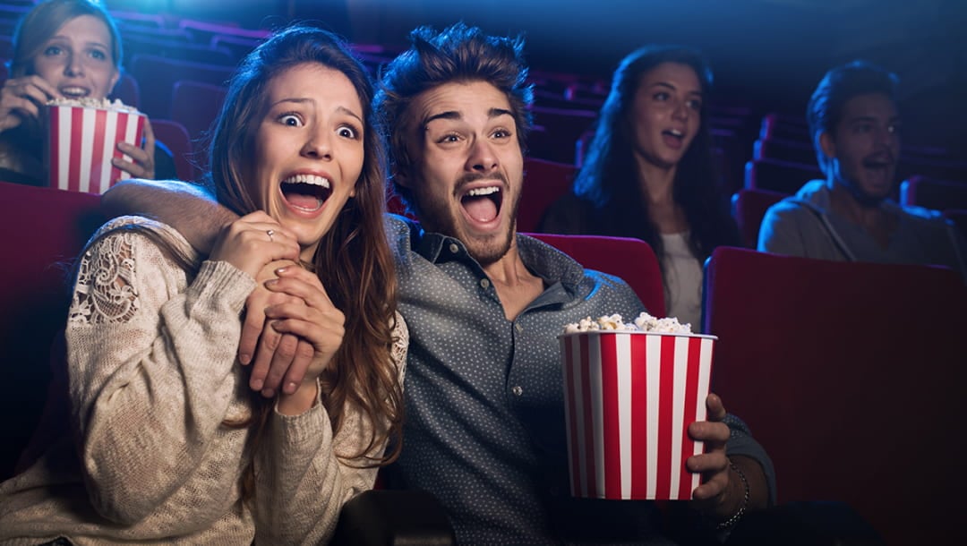 A man and woman sitting on red seats in a movie theater. The man holds a red and white striped box of popcorn in one hand and has his arm over the woman’s shoulders.