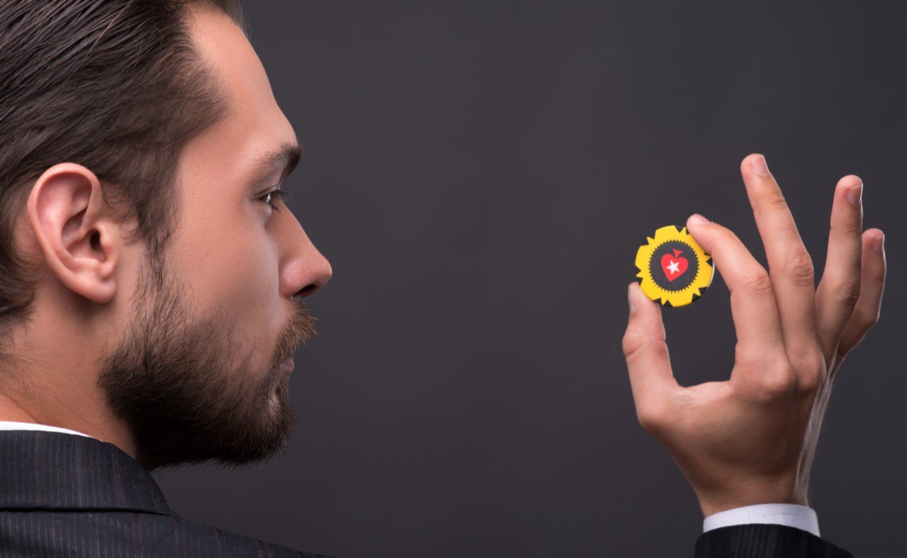 A bearded man in a suit holding up a yellow poker chip against a dark gray background.