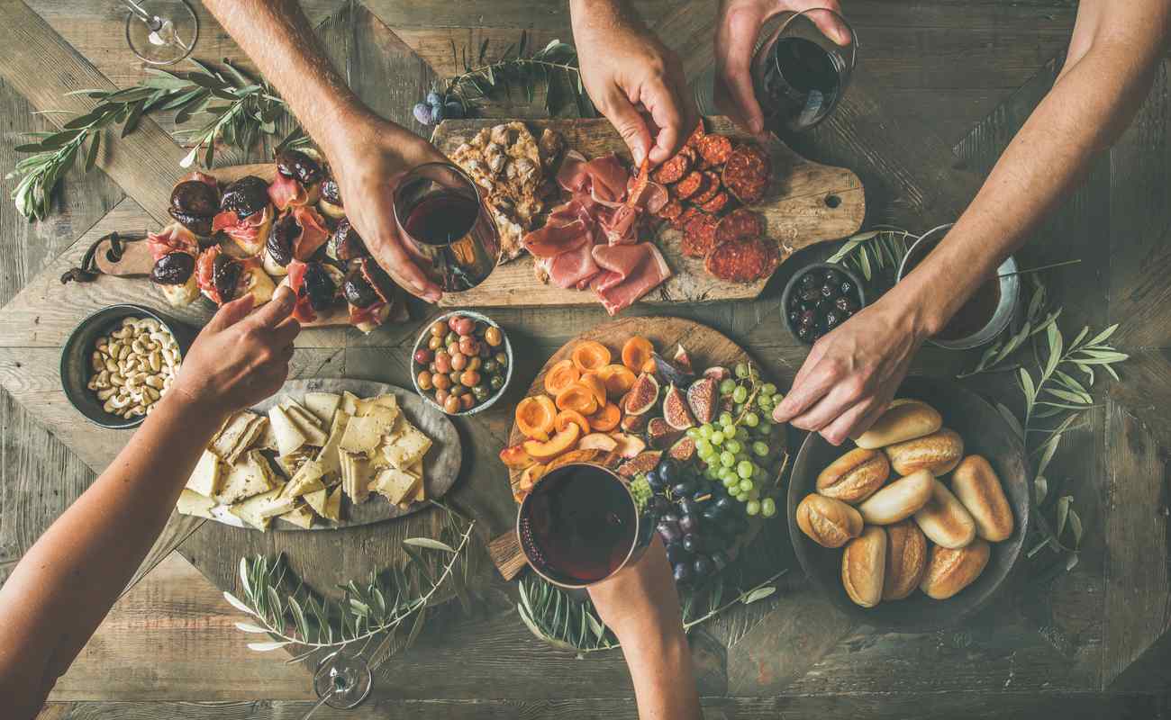 People help themselves to various snacks from a platter that includes fruit, cold meats, and cheese.