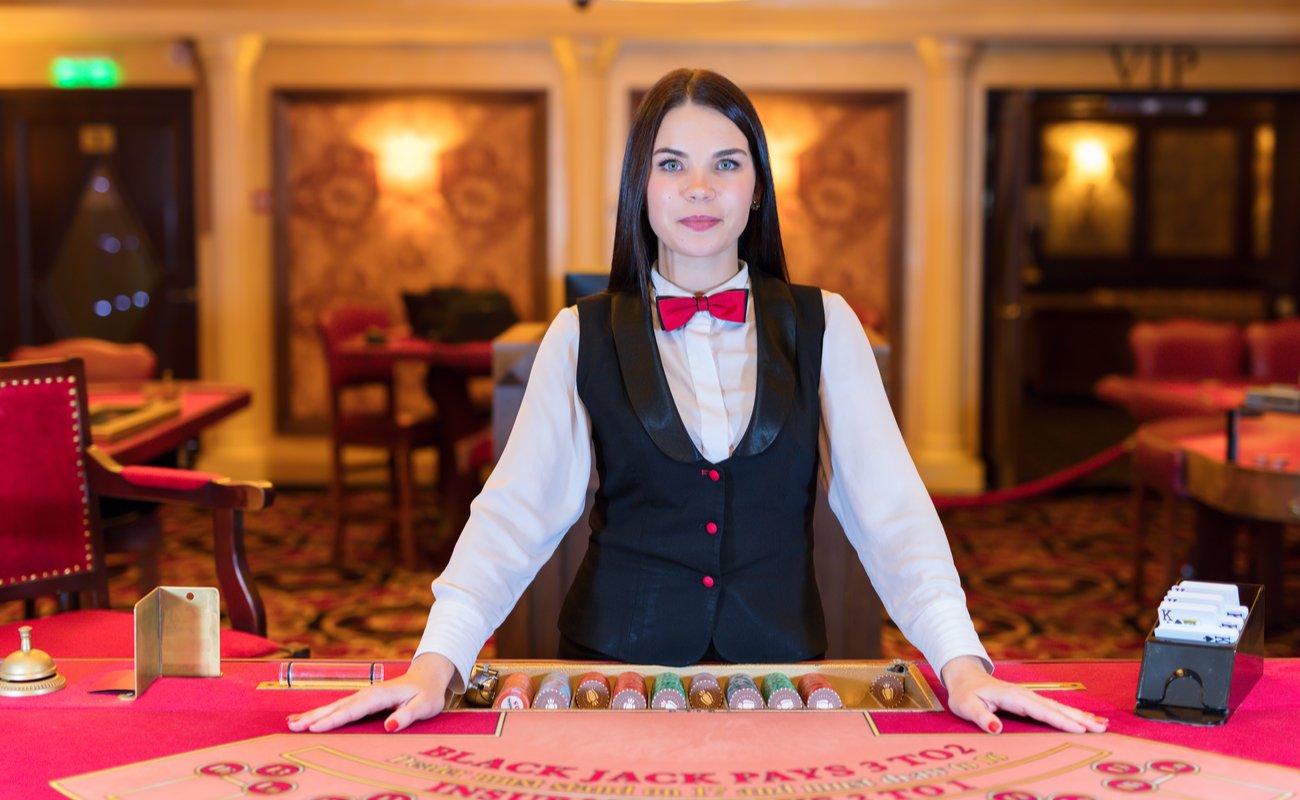 A woman dealer in white shirt, navy waistcoat and red bow tie stands at a blackjack table.