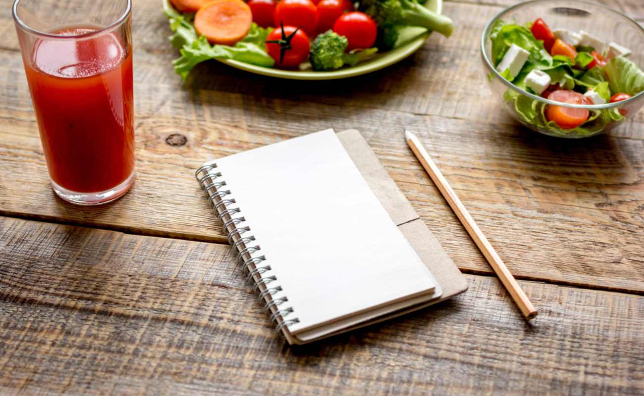 A notepad sits on a table next to a glass and some plates of food.
