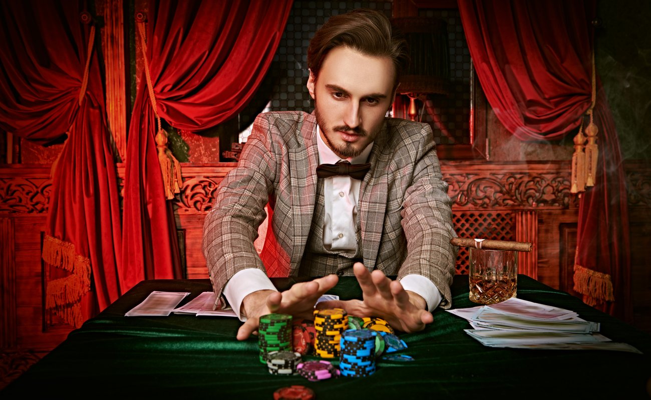 A well-dressed man pushes a pile of casino chips forward with lush red drapes in the background.