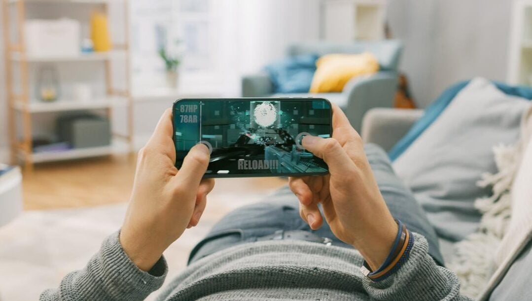 Man lying on a couch playing a video game on his smartphone with his living room in the background.