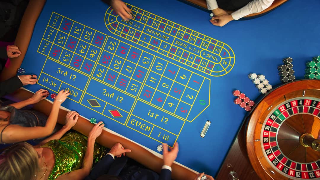 A top-down view of men and women placing bets at a roulette table. There is a roulette wheel and casino chips on the roulette table.