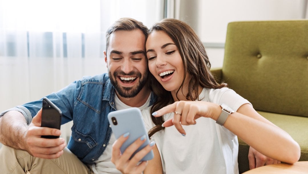 A couple sitting on the living room floor pointing at a mobile phone.