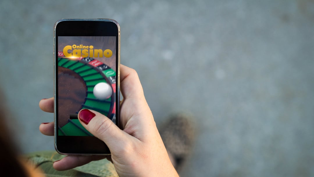 A person with painted nails holding a cellphone. The words “Online Casino” are on screen with imagery of a roulette table.