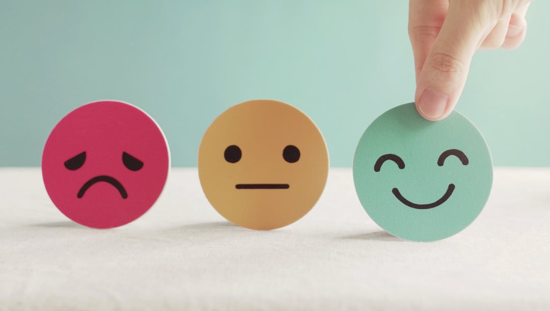 Small emoji cut-outs on top of a white surface. There’s a pink, sad emoji, a yellow, neutral-feeling emoji, and a blue, smiling emoji.