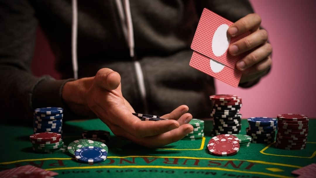 A man seated at a blackjack table, holding two cards in his hand, with casino chips placed on the table.