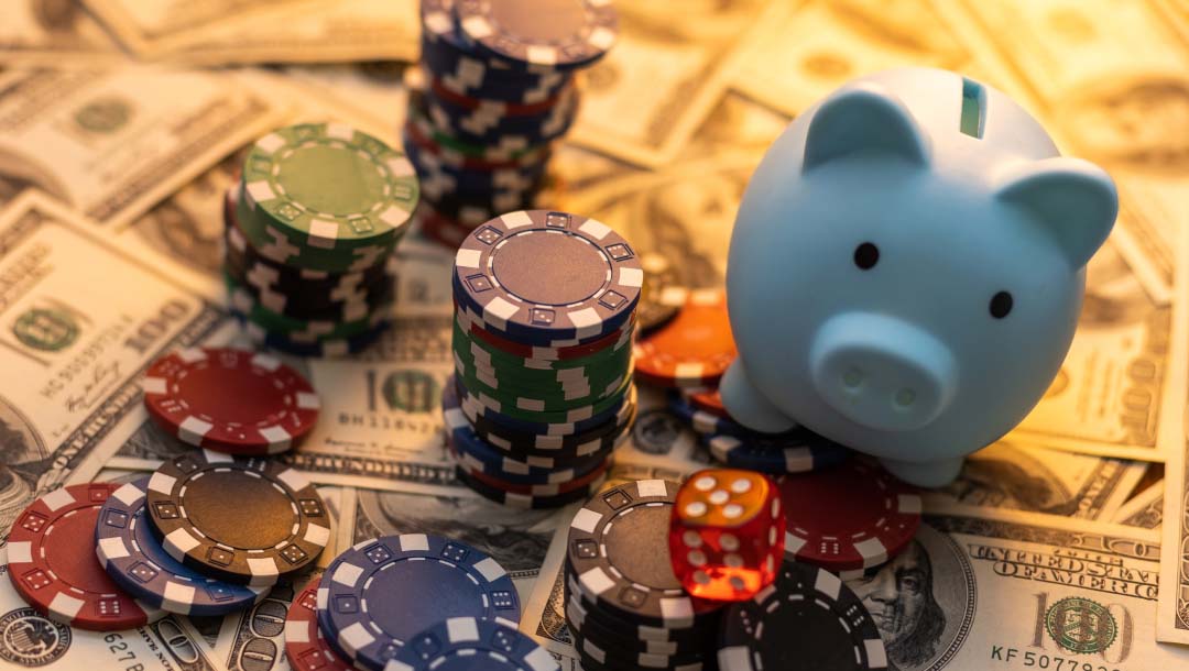 Stacks of casino chips and casino chips loosely scattered on top of $100 bills. A single dice and piggy bank are to the left of these casino chips.