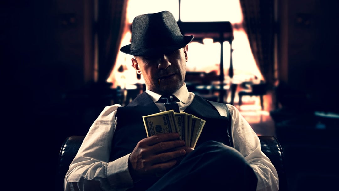 A gangster sitting on a couch in a dimly lit room counting money.