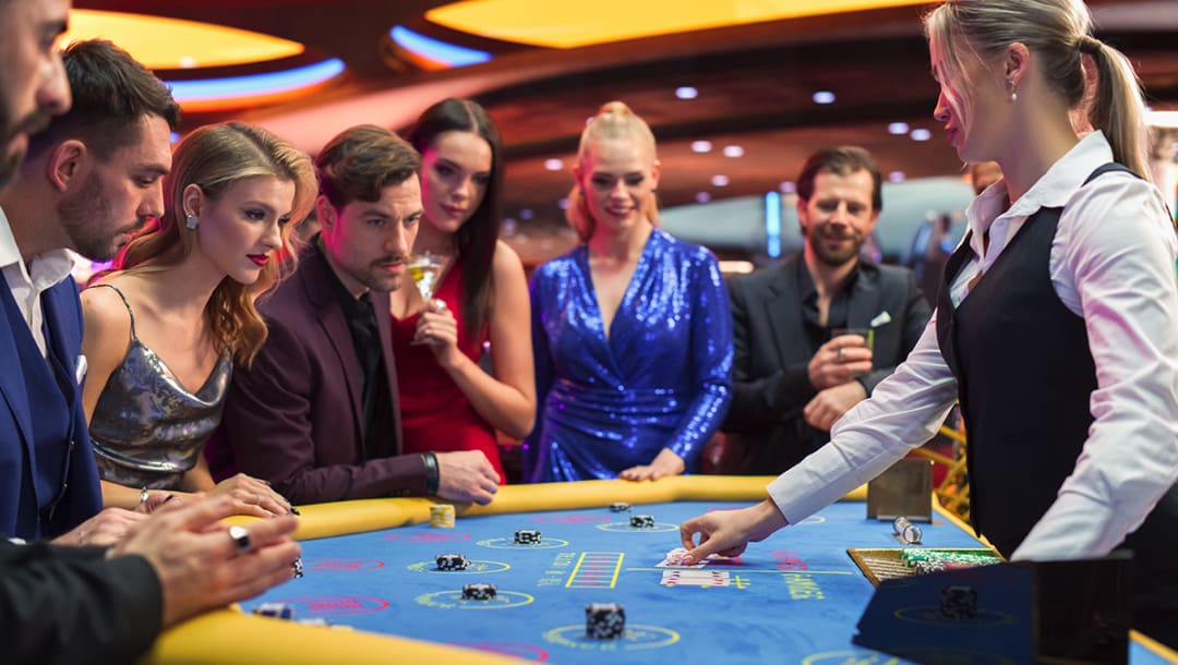 A group of people standing at a baccarat table. The baccarat dealer is touching playing cards on the blue baccarat table.