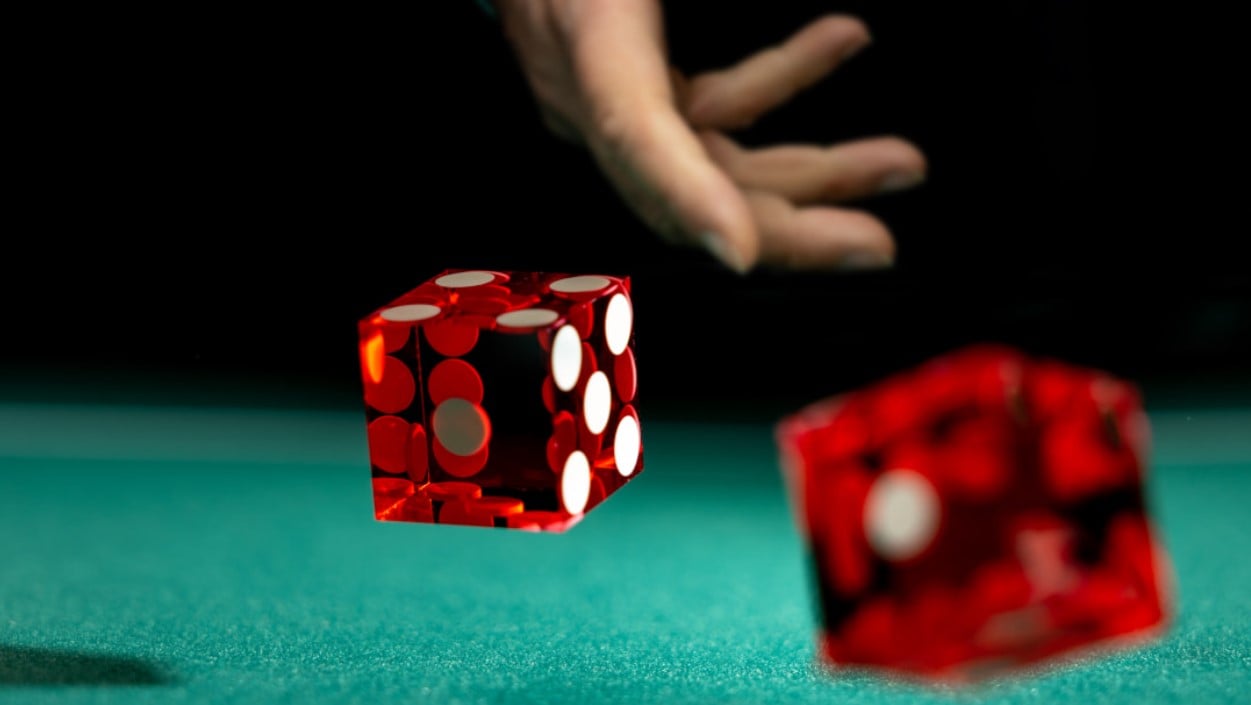 A closeup of two red dice being thrown onto a craps table. The dice are still in the air, just about to land on the green felt of the table and the player’s hand is still in the frame.