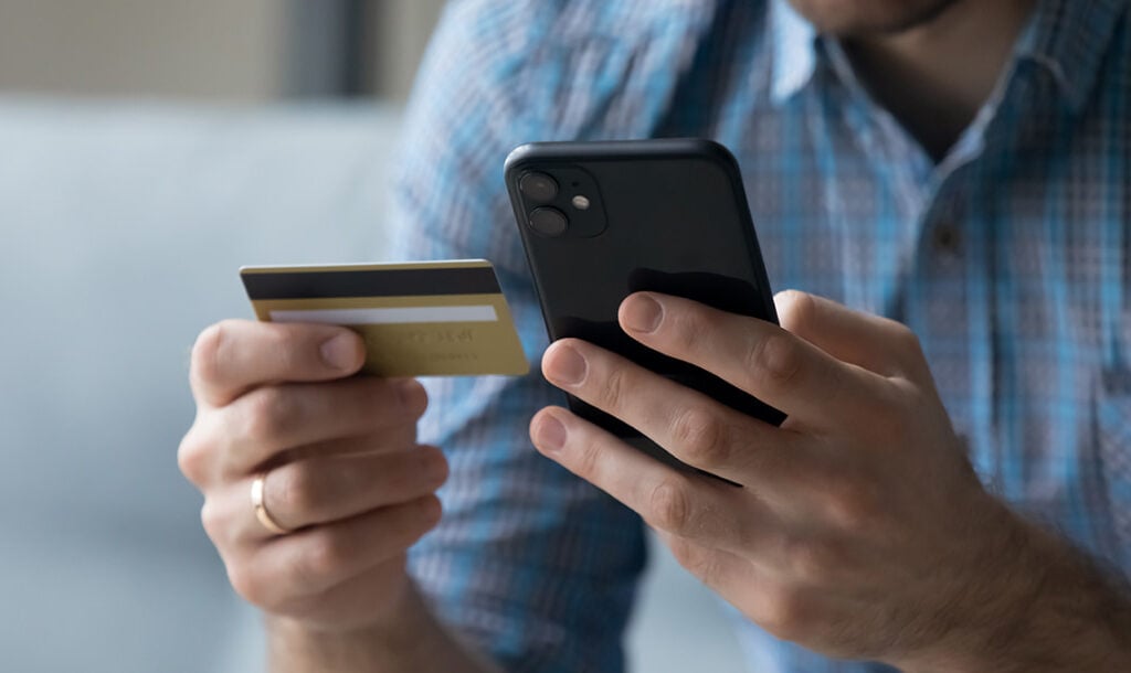 A concept image for online payments. The photograph is a closeup of a man in a button-up shirt holding a smartphone in his left hand and a bank card in his right hand.