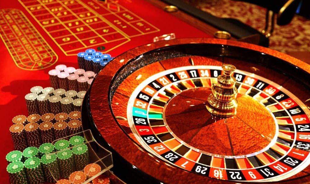 A closeup photograph of a traditional roulette table. Stacks of casino chips are neatly arranged on the red felt alongside the roulette wheel.