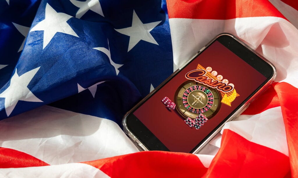 A photograph of a smartphone showing vector-style casino imagery, such as flashing signs, casino chips, and a roulette wheel. The phone is on top of an American flag, which is used as the background of the image.