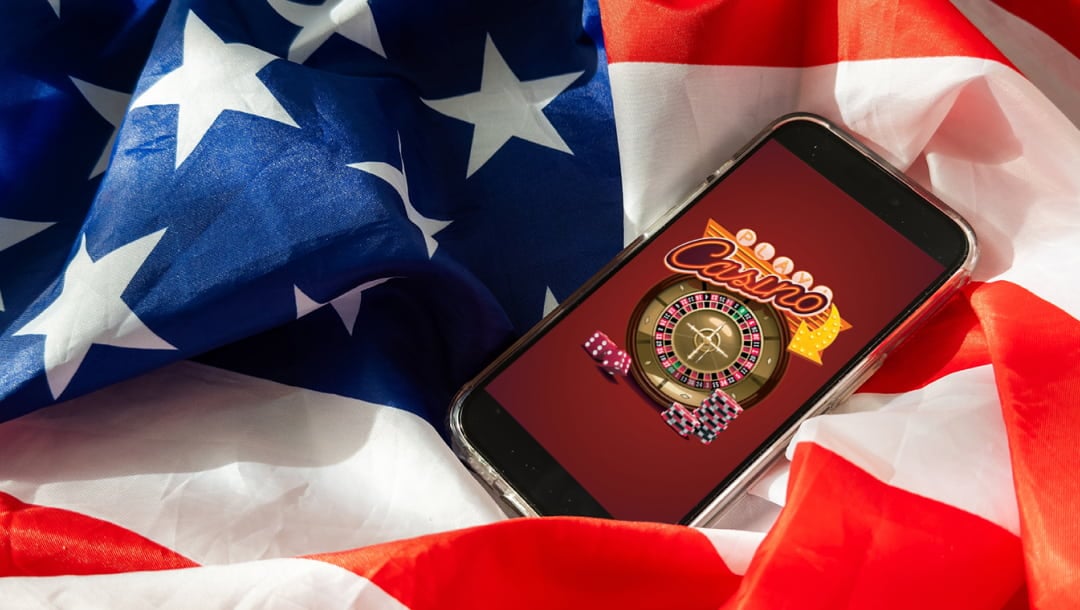 A photograph of a smartphone showing vector-style casino imagery, such as flashing signs, casino chips, and a roulette wheel. The phone is on top of an American flag, which is used as the background of the image.