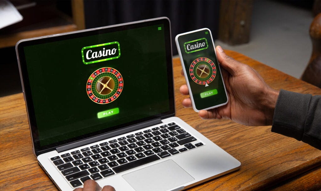 An over-the-shoulder view of a man holding a cellphone in his right hand while his left rests on the laptop in front of him. Both devices have online roulette loaded on the screen with a “play” button to start the games.